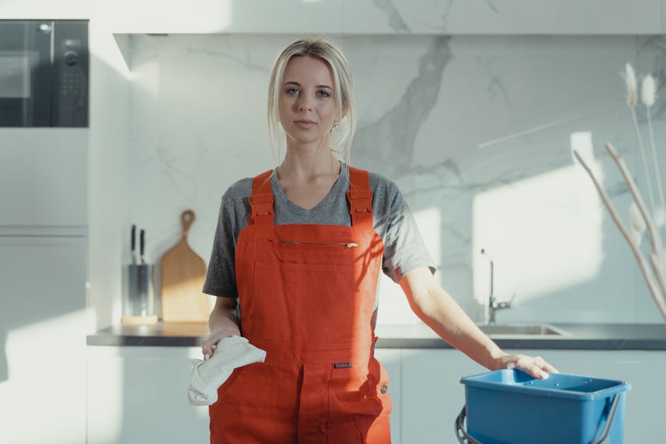 A woman wearing a red cleaning apron and grey t-shirt standing in a modern kitchen holding a white cloth in her right hand and resting her left hand on a blue mop bucket. The kitchen features a white marble backsplash, stainless steel appliances, and a wooden cutting board on the counter. Sunlight illuminates the space, highlighting the clean, shiny surfaces of the countertops and the stainless steel sink. The image emphasizes surface cleaning and hygiene as part of professional domestic cleaning services offered by Cleaners SW6, reflecting thorough and hygienic cleaning processes typically involved in deep cleaning and surface sanitisation.