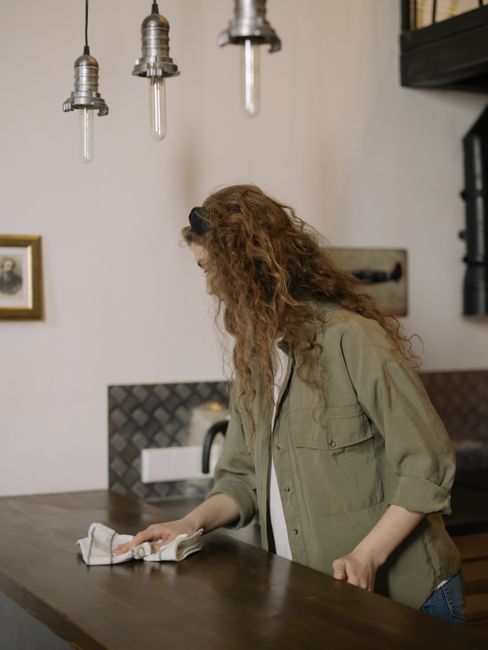 A woman with long, curly red hair wearing a beige jacket is engaged in household cleaning, using a cloth to wipe a dark wooden kitchen countertop. The scene features three industrial-style pendant lights hanging from the ceiling, which is painted in a neutral tone. In the background, there is a white wall with framed pictures and a black shelf system. The countertop appears clean and free of dust or debris, reflecting good hygiene practices. The kitchen surfaces, including the splashback and cabinetry, are well-maintained and exhibit a polished, gleaming finish. Natural lighting illuminates the space, highlighting the clarity and cleanliness of the surfaces, which are made of wood, ceramic, and stainless steel. This setting exemplifies the quality of domestic surface cleaning and sanitisation services offered by Cleaners SW6, aligning with the focus on care for period home cleaning near Fulham Palace SW6 as featured on the page.
