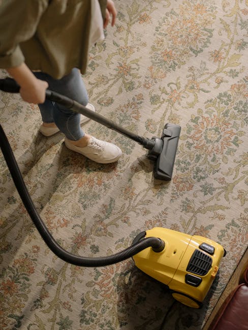 A person using a vacuum cleaner with a yellow body and black attachments to perform deep surface cleaning on a traditional patterned area rug, which features floral motifs in muted tones of beige, green, and peach. The rug is laid out on a light-colored wooden floor in a residential room. The individual is wearing casual clothing, including beige shoes and jeans, and is holding a vacuum wand that extends to the rug. The room is well-lit, highlighting the cleanliness of the rug and the equipment used by Cleaners SW6 for domestic cleaning services in Fulham, SW6, as promoted on the company’s website.