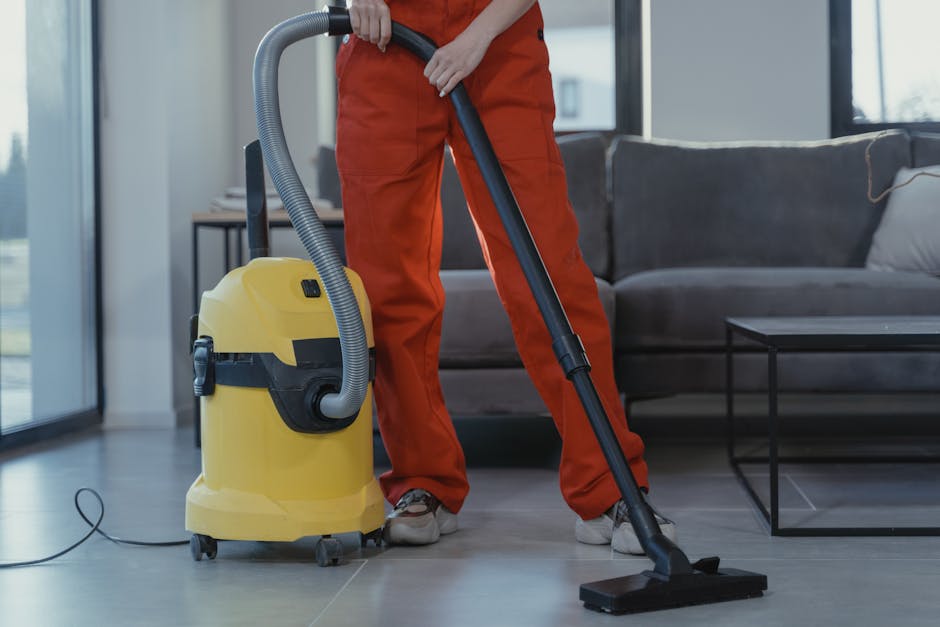 A professional cleaner from Cleaners SW6 is using a yellow vacuum cleaner with a black hose to perform surface cleaning on the light grey tiled floor of a living room in a residential setting. The cleaner, dressed in orange work overalls and grey shoes, is holding the vacuum hose with both hands, directing it across the floor near a black metal coffee table. The room features a dark grey sofa with light-colored cushions, large windows allowing natural light, and a modern interior design with a minimalist aesthetic. The polished tiles appear clean and dry, reflecting the natural light from the windows, indicating a thorough deep cleaning process.