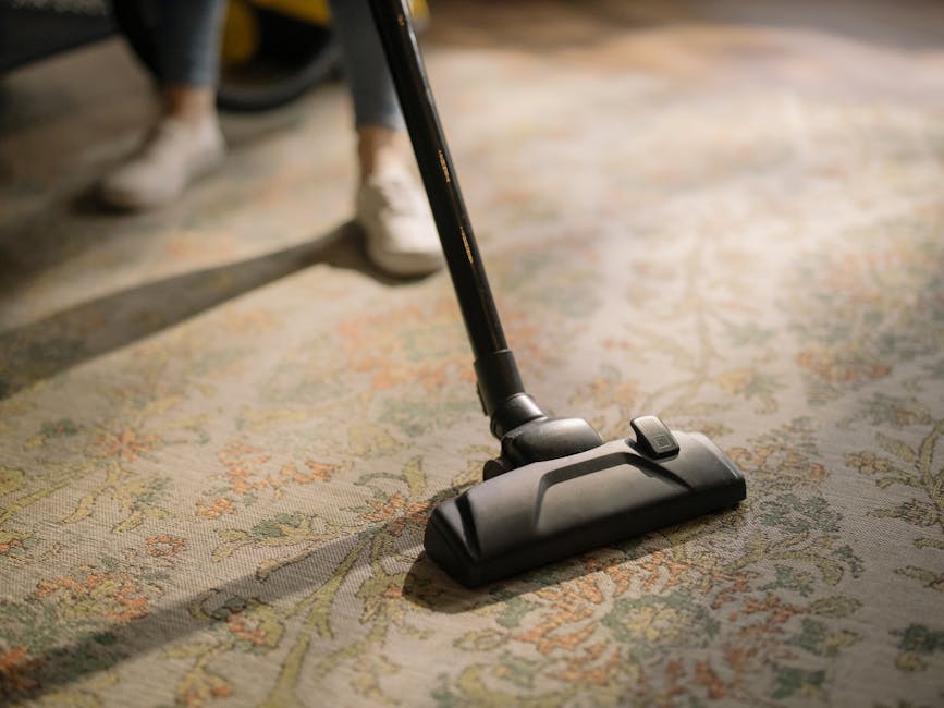 A person using a vacuum cleaner with a yellow body and black attachments to perform deep surface cleaning on a traditional patterned area rug, which features floral motifs in muted tones of beige, green, and peach. The rug is laid out on a light-colored wooden floor in a residential room. The individual is wearing casual clothing, including beige shoes and jeans, and is holding a vacuum wand that extends to the rug. The room is well-lit, highlighting the cleanliness of the rug and the equipment used by Cleaners SW6 for domestic cleaning services in Fulham, SW6, as promoted on the company’s website.