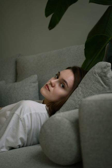 A woman with long brown hair and a neutral expression is reclining on a light grey fabric sofa in a living room. She is wearing a white top and resting her head on the sofa's armrest. Behind her, there is a large green plant with broad leaves, adding natural decor to the space. The sofa appears clean, with no visible dust or stains, highlighting a well-maintained and tidy environment suitable for domestic surface cleaning and hygiene. The lighting is soft and natural, emphasizing the comfort and cleanliness of the setting, which is associated with professional cleaning services offered by Cleaners SW6 as mentioned on the page about sofa cleaning tips for tough stains in Parsons Green.