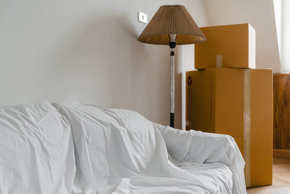 A living room scene featuring a white sofa with a slipcover that appears slightly wrinkled, situated against a plain beige wall. To the right of the sofa, there are three cardboard moving boxes stacked in descending size order, with the top box slightly tilted. A tall, standing floor lamp with a brown, conical fabric shade is positioned behind the boxes, casting soft light onto the area. The room is illuminated by natural light coming from an unseen window on the right, creating a bright and tidy environment. The floor appears to be wooden, and the overall setting suggests a space prepared for deep cleaning or maintenance, with a focus on surface cleanliness and organisation, as referenced in the Parsons Green sofa cleaning insider tips for tough stains for SW6 by Cleaners SW6.