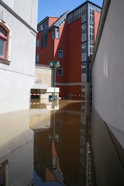 A flooded urban alleyway with water partially submerging the pavement, reflecting nearby buildings including a modern red-brick apartment complex with large glass windows and a grey and white neighboring structure. A streetlamp with a green pole and double light fixtures stands amidst the floodwaters. The scene is illuminated by natural daylight, showcasing the clean, wet surfaces of the alley. The image emphasizes the importance of surface cleaning and sanitation in flood-affected areas, aligning with the services offered by Cleaners SW6 in Fulham for urgent flood cleaning help. The reflections in the water highlight the clarity and condition of the surfaces, demonstrating the need for professional deep cleaning and sanitisation to restore a safe, hygienic environment.