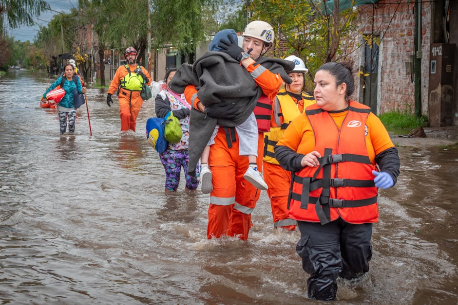 A group of rescue workers in bright orange and red waterproof jackets and trousers wade through a flooded street with water reaching their knees. One rescuer is carrying a young child wearing a grey hoodie and white sneakers, while others follow behind with backpacks and safety gear. The street is lined with trees with green leaves and buildings with brick facades, some with visible flood damage. The scene depicts emergency response efforts during flooding, highlighting the need for professional flood cleaning and recovery services, as provided by Cleaners SW6, to restore safety and hygiene to affected homes and businesses in Fulham.