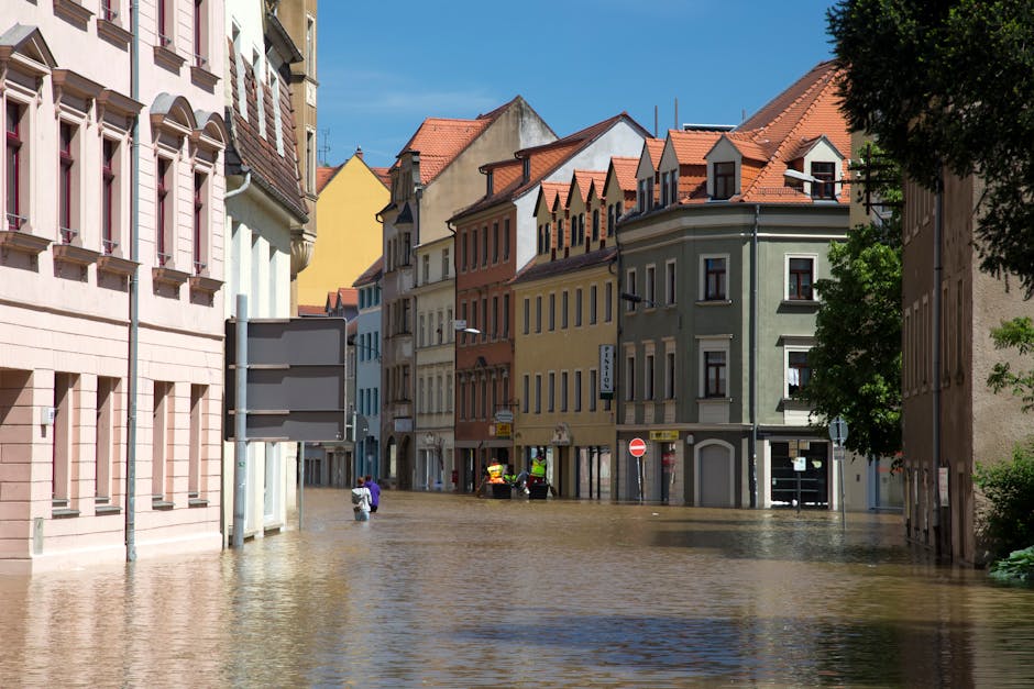 A flooded urban alleyway with water partially submerging the pavement, reflecting nearby buildings including a modern red-brick apartment complex with large glass windows and a grey and white neighboring structure. A streetlamp with a green pole and double light fixtures stands amidst the floodwaters. The scene is illuminated by natural daylight, showcasing the clean, wet surfaces of the alley. The image emphasizes the importance of surface cleaning and sanitation in flood-affected areas, aligning with the services offered by Cleaners SW6 in Fulham for urgent flood cleaning help. The reflections in the water highlight the clarity and condition of the surfaces, demonstrating the need for professional deep cleaning and sanitisation to restore a safe, hygienic environment.